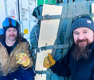 Two men crouching beside a large, cloth-covered parcel bound with several straps, giving thumbs-up gestures to the camera. A plank of wood strapped against the parcel bears a handwritten message that reads: "Hey Antarctica! Hope this gets to you safe. From the guys at 176!" The message is followed by a list of names