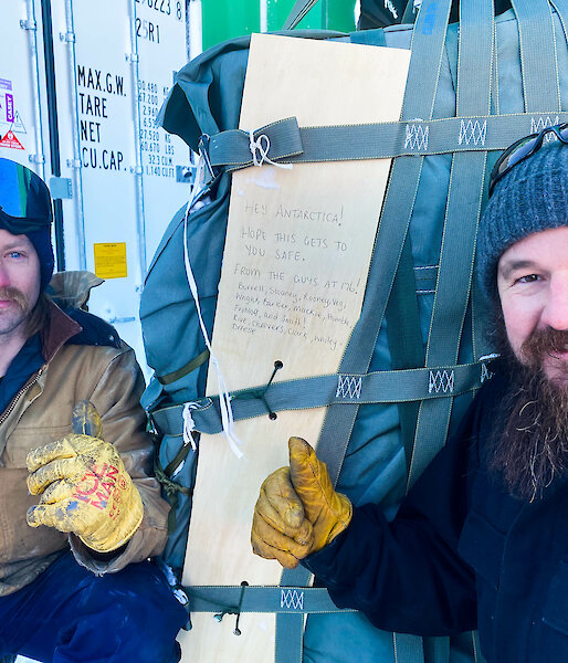 Two men crouching beside a large, cloth-covered parcel bound with several straps, giving thumbs-up gestures to the camera. A plank of wood strapped against the parcel bears a handwritten message that reads: "Hey Antarctica! Hope this gets to you safe. From the guys at 176!" The message is followed by a list of names