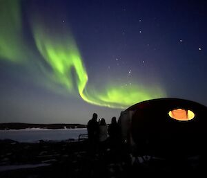 Group of expeditioners stand over front porch of field googie, looking at aurora in the distance