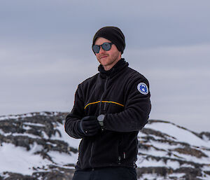 A portrait-like photo of a young man in front of a snow-laden hill in Antarctica. He is wearing a black polarfleece top, a black beanie and gloves, and round sunglasses