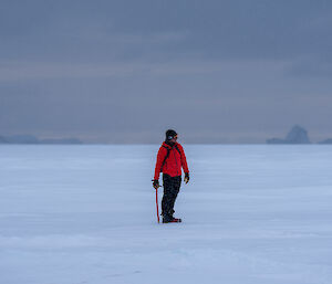 A man in a bright red jacket, carrying a red-handled ice axe, standing on a flat stretch of sea ice. Icebergs can be seen in the distance against a sky dark with snow clouds