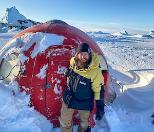 A man in a black beanie stands in the snow in front of a hut