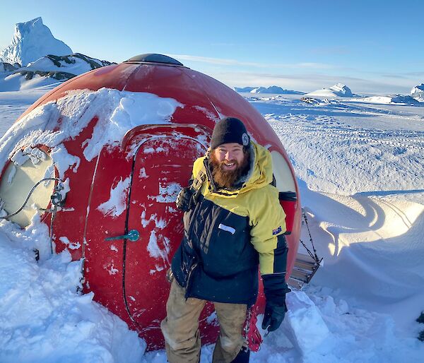 A man in a black beanie stands in the snow in front of a hut