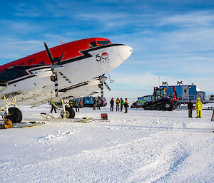 A Basler plane with skis attached to its wheels, parked on a snowy plain. An engineer is doing some work on on of the wheel-ski assemblies. Groups of people stand in the background alongside a brightly-painted tractor, a light blue shed mounted on a sled and a couple of Hägglund vehicles