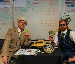 Two men, sprucely dressed in waistcoats, ties, smart jackets and hats, sit at a small square table in front of a whiteboard, holding wads of cash in their hands. Names of Melbourne Cup entrants and betting odds are written on the whiteboard