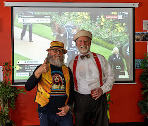 Two men standing in front of a projector screen on which the Melbourne Cup horse race is being shown. One man is wearing a yellow-gold waistcoat and hat and giving a thumbs-up gesture to the camera. The other man wears bright red suspenders and a bow tie with a white shirt