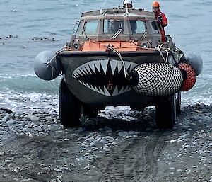 A vehicle capable of water and land transport drives out of the ocean and up a grey sandy beach