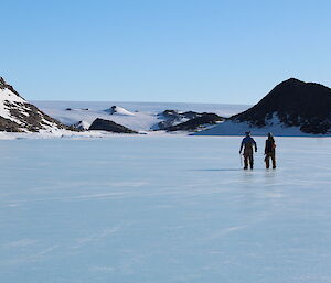 Two people walk across an icy surface under a blue sky