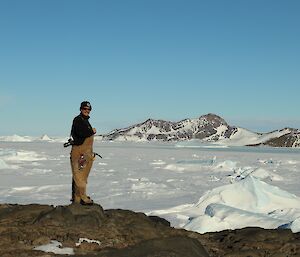 A man stands on a rocky outcrop with a view of snow and  mountains