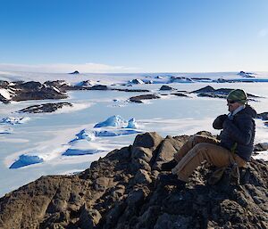 A person sits on a rocky outcrop looking at a vast snowy view