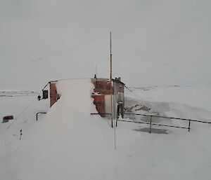 A small wooden hut in a snowy field. A large bank of snow has built up against one of its walls. An amateur radio antenna, constructed of a slim pole a few metres tall with a couple of long wires fanned out at the top, has been planted in the snow just outside the hut