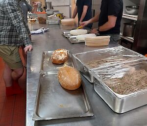 A long stainless steel kitchen table with stacks of proving baskets and a couple of loaves of freshly baked bread. A group of people stand around the table, watching a chef preparing more loaves to go in the oven