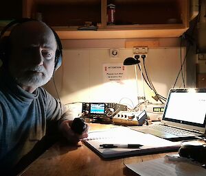 A man with headphones on, sitting at a desk in a dim room lit only by a desk lamp. On the desk in front of him are a laptop computer, notebook pen, and two small radio units with tuning knobs and a display screen