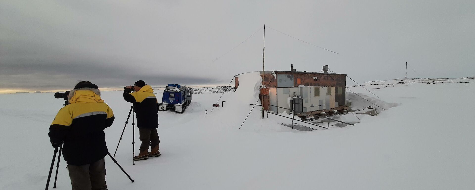 A small wooden hut in a snowy field. Outside the hut, two people in thick winter jackets are peering through cameras set up on tripods