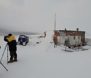 A small wooden hut in a snowy field. Outside the hut, two people in thick winter jackets are peering through cameras set up on tripods