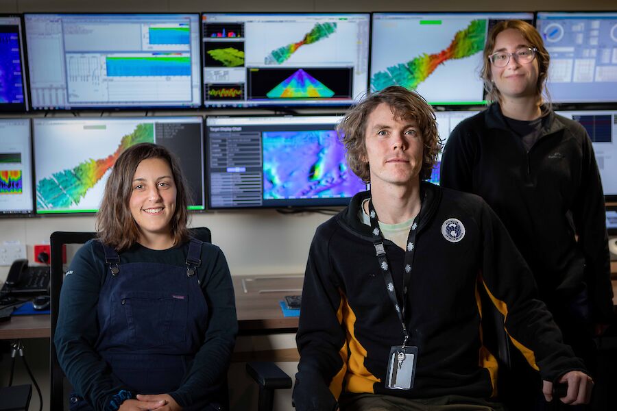 3 people in front of a wall of colourful seafloor monitors smiling to camera