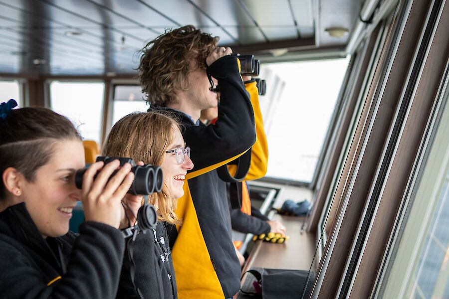 Three people, two with binoculars up, stand at the windows on the viewing deck looking out