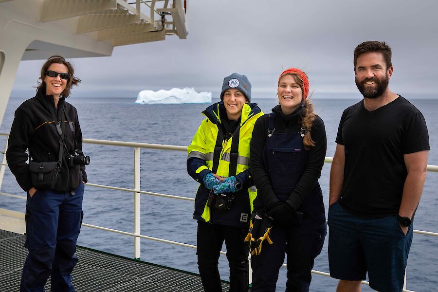 4 expeditioners pose on deck with big smiles, with an iceberg in the background