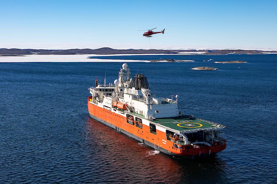 An aerial of a ship with a helicpoter above, about to fly-off a load of cargo