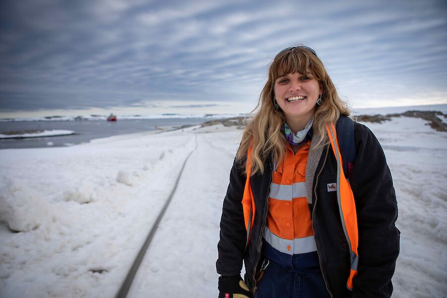 A woman smiles to camera while standing beside a long length of fuel line in the snow