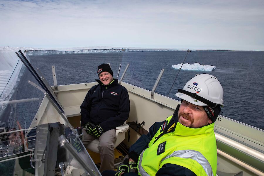 Two men in a box at the back of a ship smiling to camera.  A large glacier is behind them