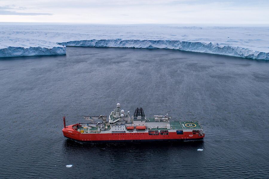 An aerial of a red research ship sitting just offshore from a large glacier