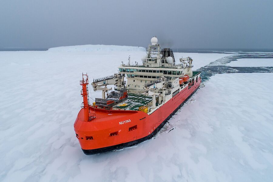An aerial of a large red and white boat as it cuts a path through some soft ice heading towards camera