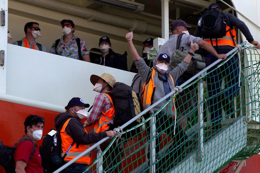 A queue of people on a gangway waiting to board a ship.  One man stands with arms aloft facing the camera