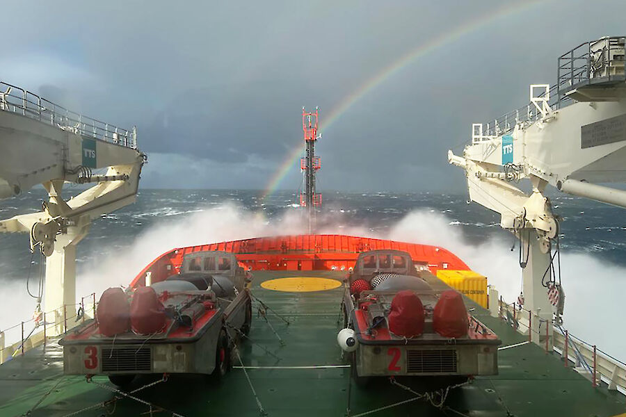 The bow of a ship with waves breaking over the front.  A rainbow is in the sky just ahead.