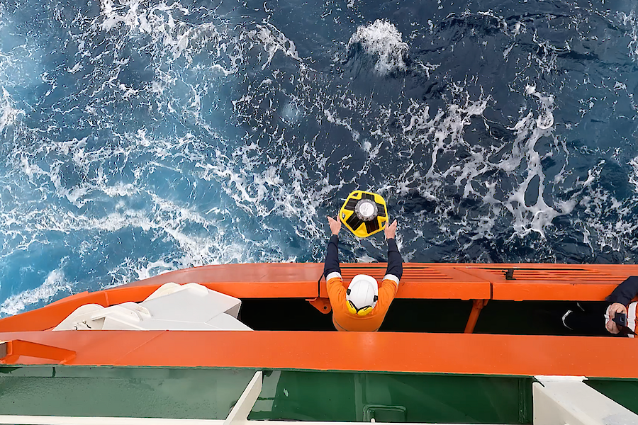 Looking down the side of a ship from above where a man on the deck below can be seen leaning over the edge with a buoy in his hands, ready to release in to the ocean.