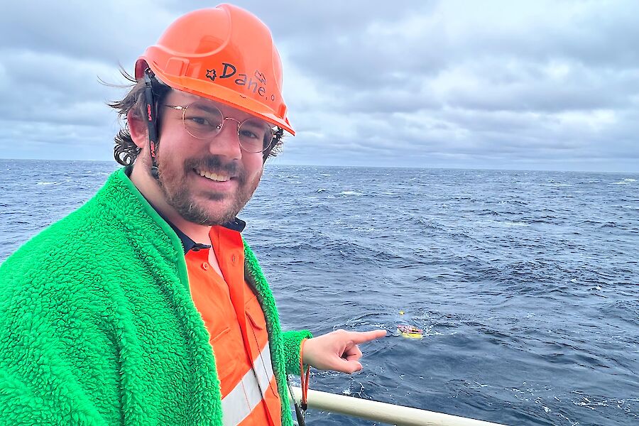 A man in a hard had and hi-vis stands at the side of a ship and points to an object in the water.  He has Dane written on his hat.