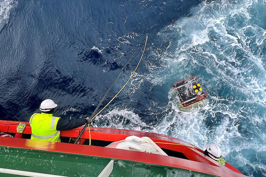 A man leans out of the back of a ship and pulls on a rope attached to an object in the water