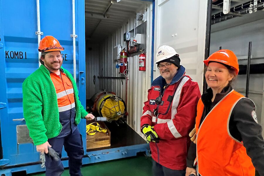3 people smile to camera beside an open shipping container.  A yellow cylindrical whale mooring is stored inside the container.