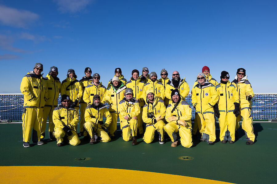 A group of people gather for a group photo on the back deck of a ship.  They are all wearing bright yellow windproof gear.