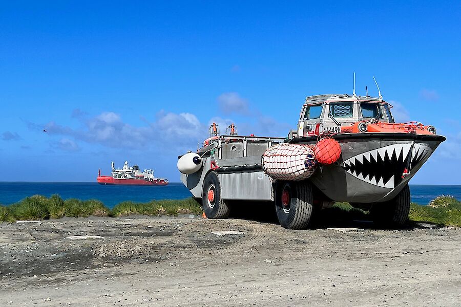 An amphibious vehicle with a large painted mouth with pointy teeth, sits on shore with a large ship visible in the sea behind