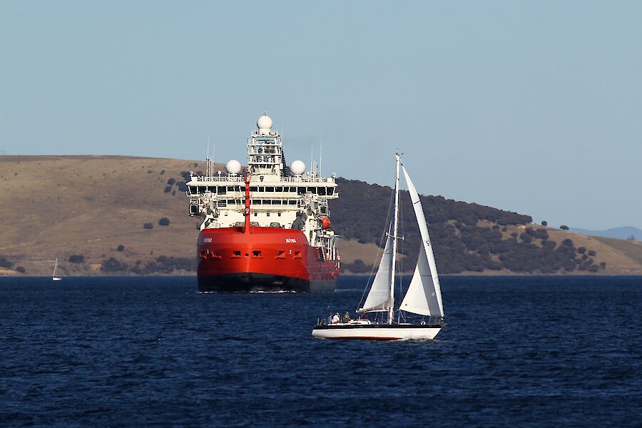 A large red ship sails towards camera with a small white sail boat tacking in front of it