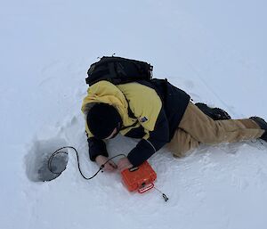 A man in a thick winter jacket and balaclava is lying on the sea ice connecting a cable to a hydrophone, a device enclosed in a case which looks like an orange lunchbox. The other end of the cable goes down through a hole drilled into the ice next to him