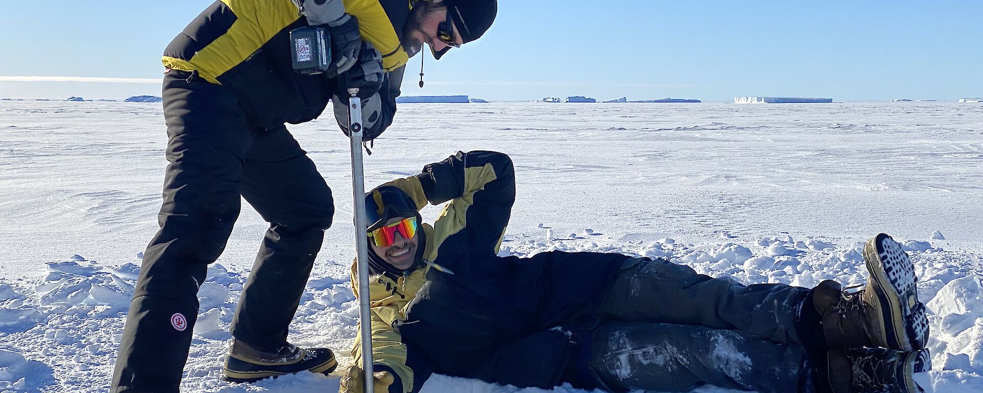 A man drilling into sea ice, leaning his weight onto a hollow drill bit over a metre long stuck perpendicularly into the ice. Another man is stretched out on his side beside the drilling hole, pretending to hold the bit steady and smiling at the camera