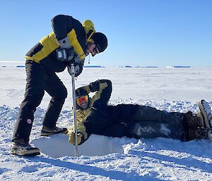 A man drilling into sea ice, leaning his weight onto a hollow drill bit over a metre long stuck perpendicularly into the ice. Another man is stretched out on his side beside the drilling hole, pretending to hold the bit steady and smiling at the camera