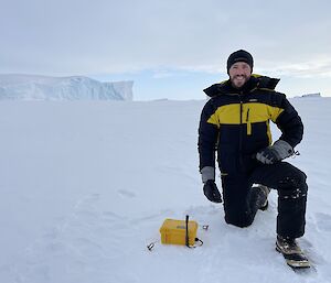 A man in a thick winter jacket and gloves, kneeling on one knee beside an ice motion buoy on a vast stretch of sea ice. The buoy is enclosed in a case that looks like a yellow lunchbox with a small antenna attached. In the near distance to the left, part of an iceberg emerging from the sea ice can be seen, looking like a blue-white, steep-edged cliff rising from the flat, white ground