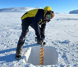 A man drilling into sea ice, legs braced wide as he leans onto the drill bit to drive it through the ice. He has turned his face up to smile at the camera. In the distance behind him, the tops of icebergs can be seen emerging from the sea ice, looking like dunes in a white desert