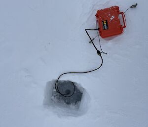 A hydrophone enclosed in an orange 'lunchbox' case lying on the sea ice. A black data cable plugged into the rear of the case unwinds down into a small, deep hole cut into the ice, into the water beneath