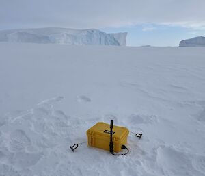 An ice motion buoy on a vast stretch of sea ice. The buoy is enclosed in a case that looks like a yellow lunchbox with a small antenna attached. In the distance, icebergs can be seen emerging from the sea ice, looking like rugged, blue-white cliffs rising from a white plain