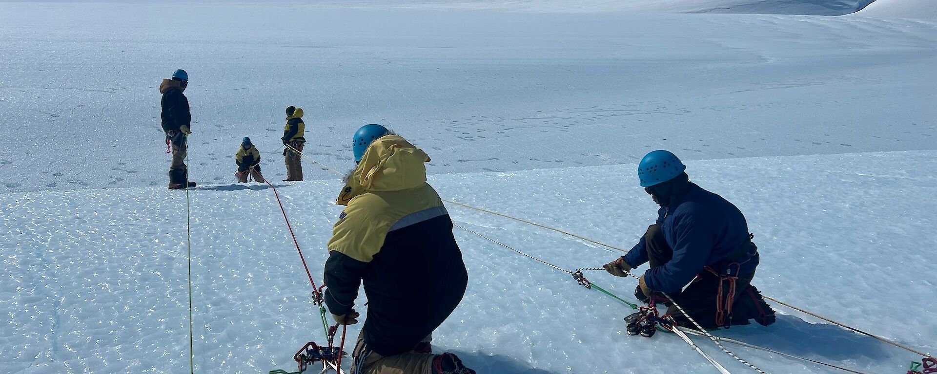 Man abseiling over ice edge with two people at top managing lines held in place on the ice