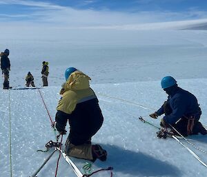 Man abseiling over ice edge with two people at top managing lines held in place on the ice