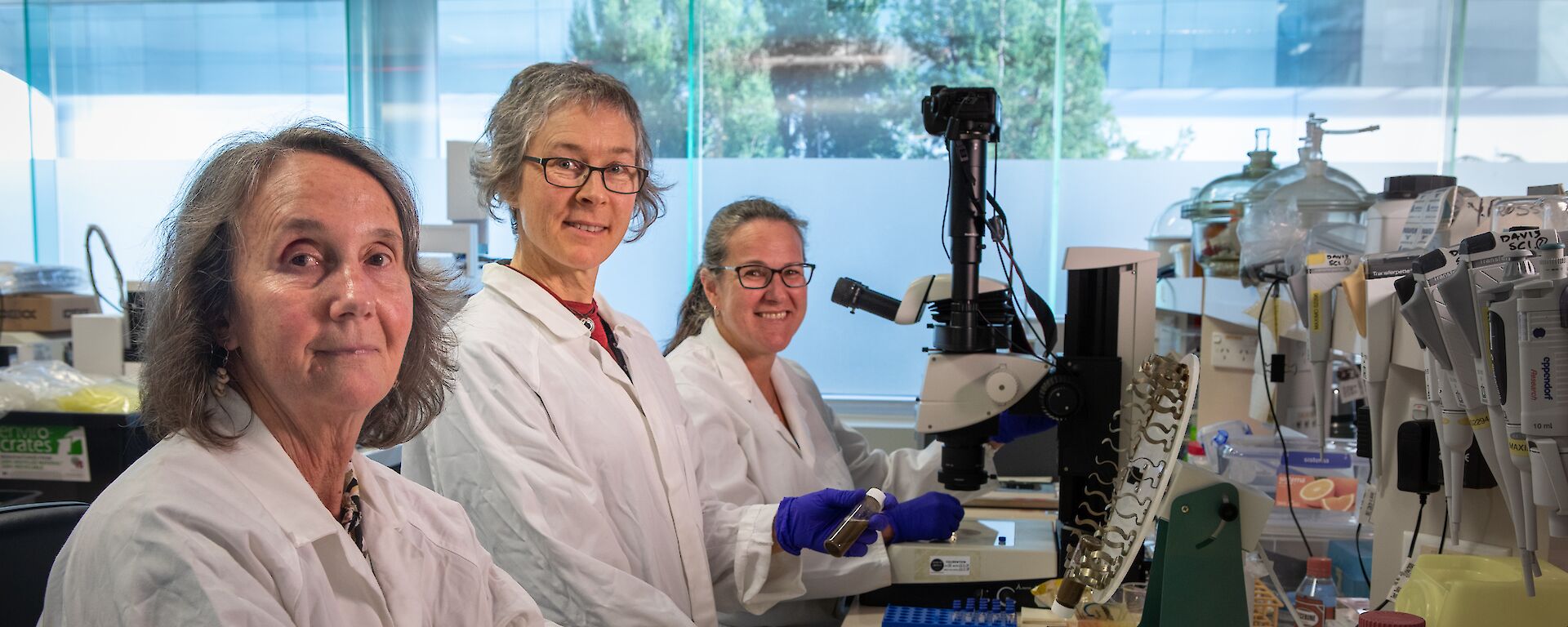Three women scientists in lab coats at a research bench.