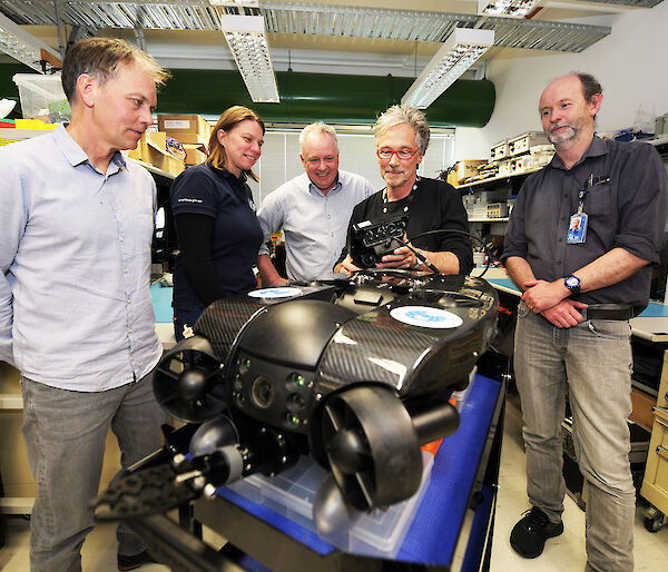 Five people gathered around a remotely operated vehicle in an electronics laboratory.
