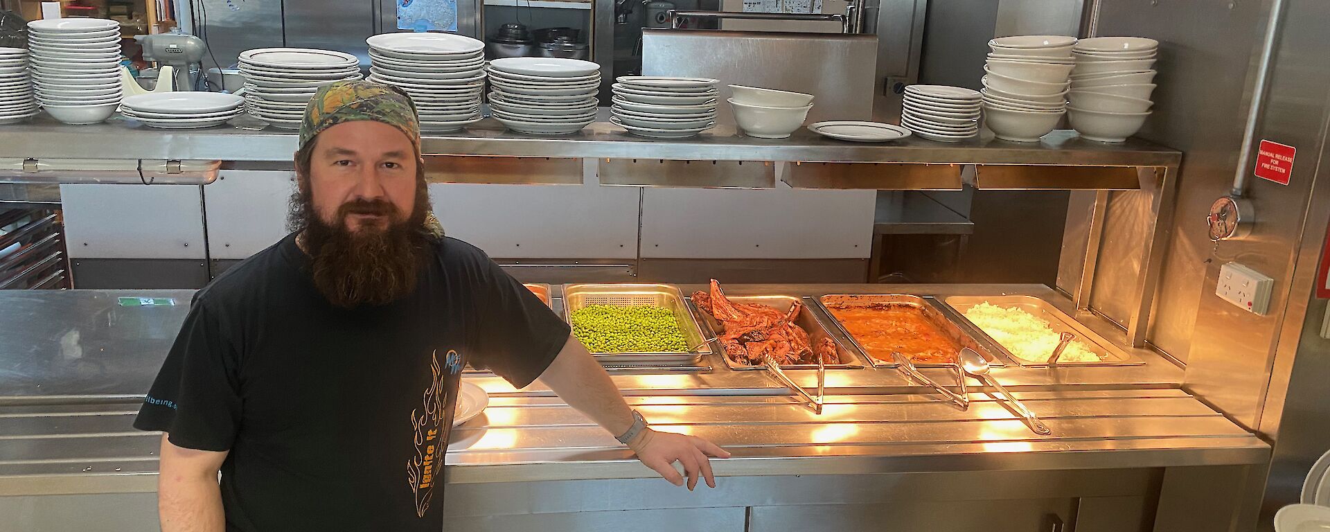 A man wearing a bandana standing in front of a stainless steel self-serve counter, with bain-maries filled with meat and vegetables he has prepared