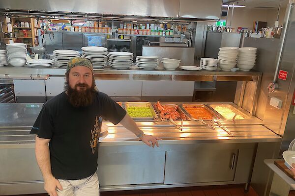 A man wearing a bandana standing in front of a stainless steel self-serve counter, with bain-maries filled with meat and vegetables he has prepared