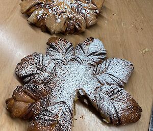 A wooden serving board with two pastries shaped like stars or snowflakes, dusted with icing sugar